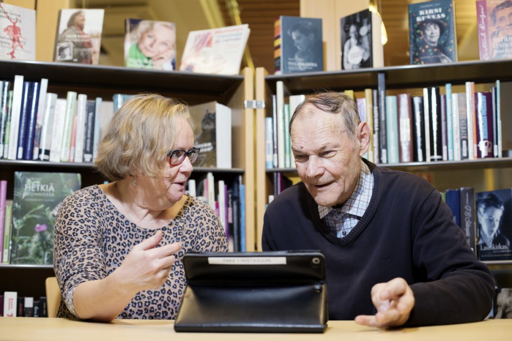 Iäkäs nainen opastaa iäkästä miestä ipadin käytössä kirjastossa.